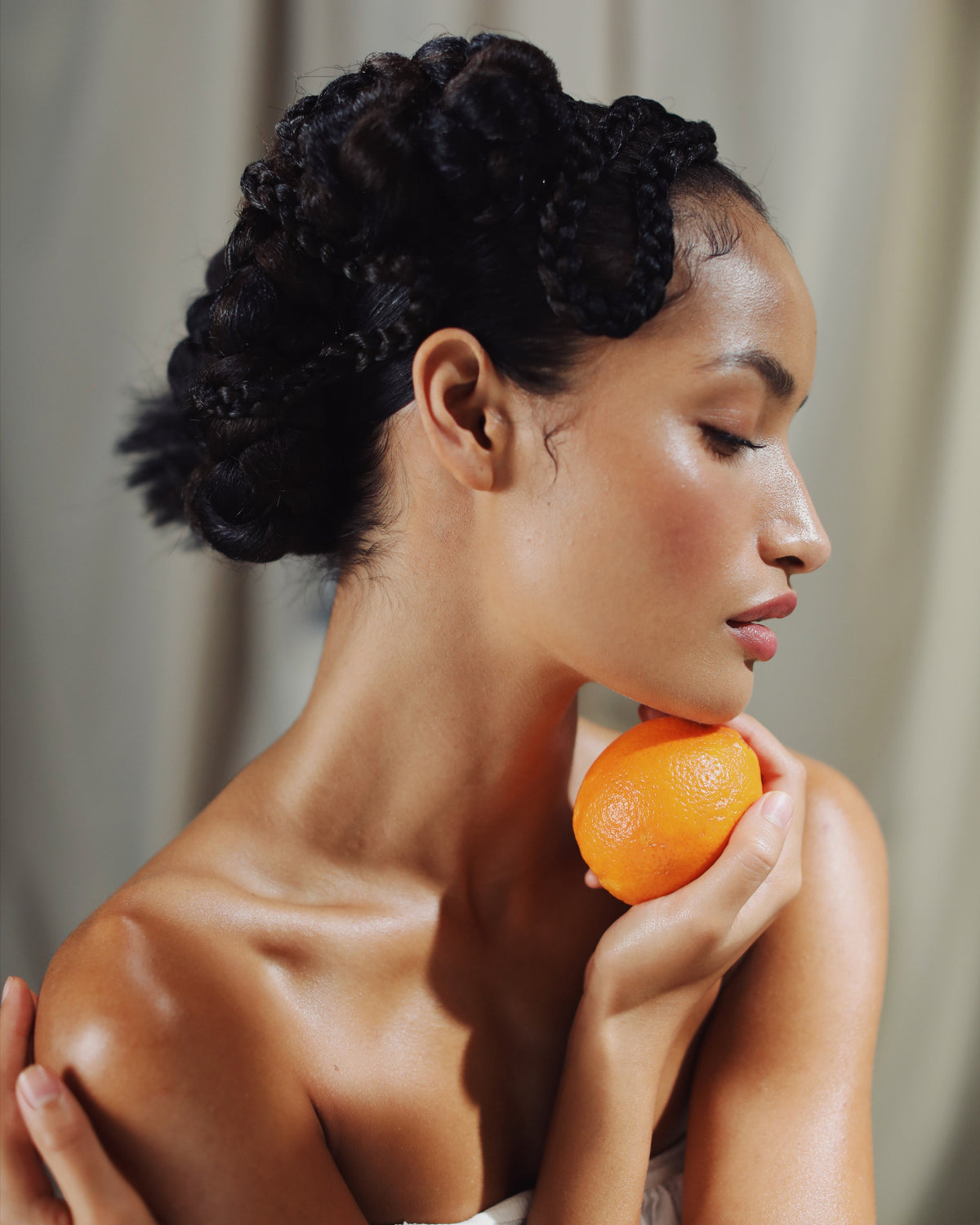 A woman with dark curly hair styled in an updo, holding an orange near her neck against a soft beige curtain background.