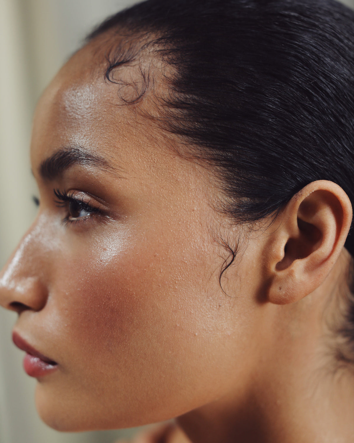Close-up side profile of a woman with dark hair pulled back, showing her forehead, ear, and neck against a soft neutral background.