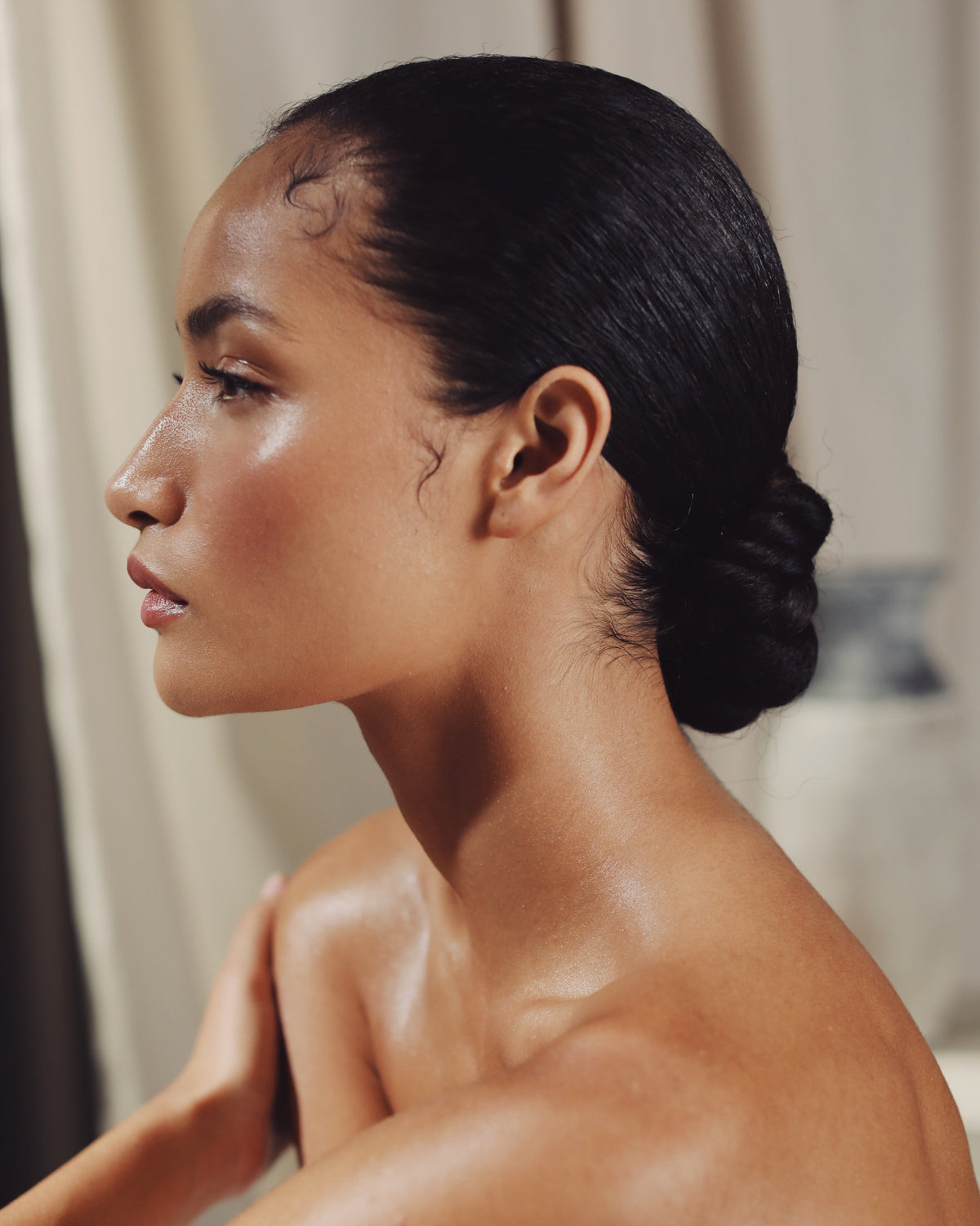 A close-up side profile of a woman with dark hair pulled back, showing her shoulder and upper chest with a glossy, moisturized finish against a soft beige curtain background.