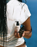 A person in a white t-shirt holds two hair care pump bottles against a solid blue background. The bottles are from the brand 'bar bur', with one in frosted white and one in dark brown.