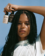 A woman with long black braids holds two bottles of bar bur cosmetic products above her head against a clear blue sky. She is wearing a white draped top.