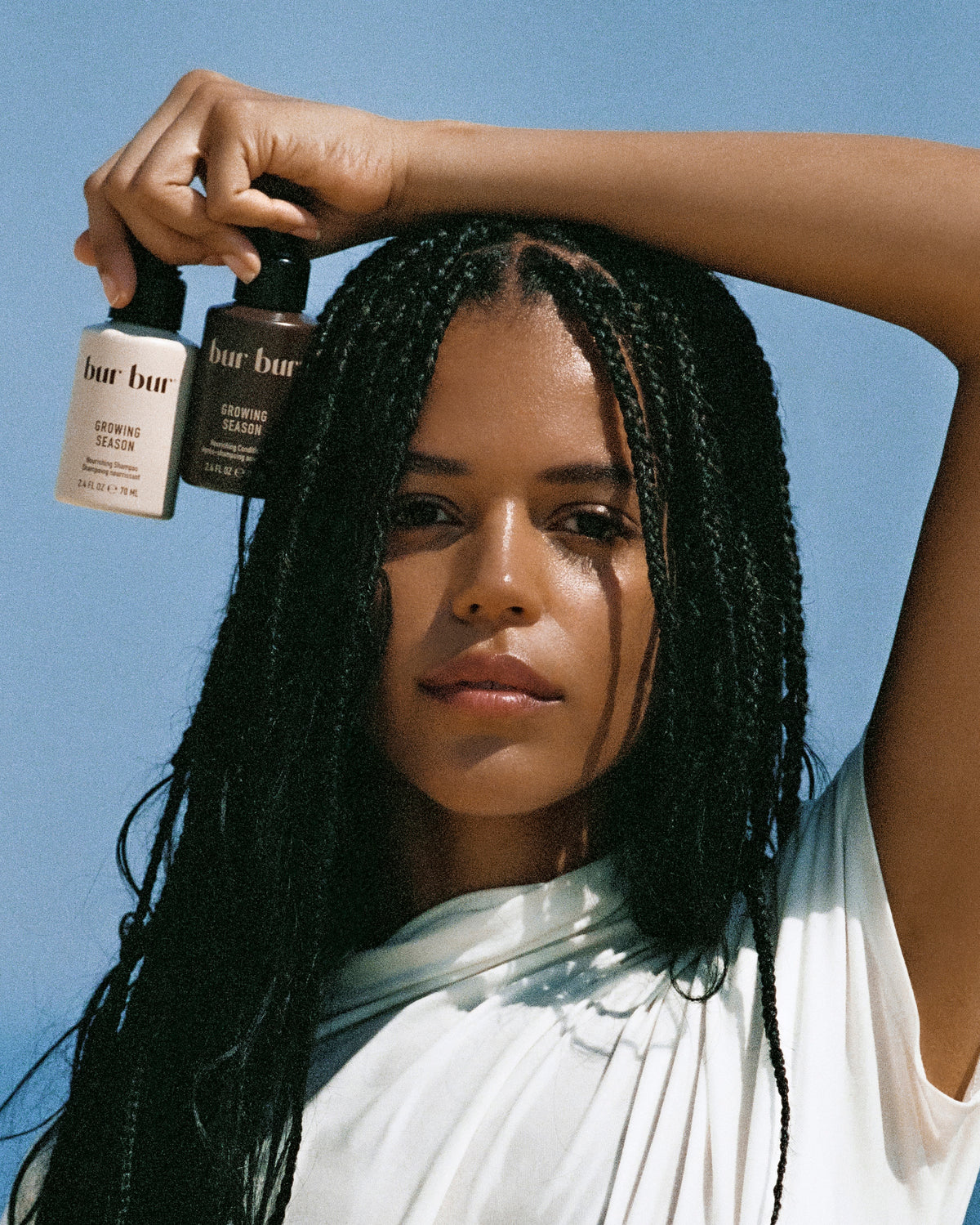 A woman with long black braids holds two bottles of bar bur cosmetic products above her head against a clear blue sky. She is wearing a white draped top.
