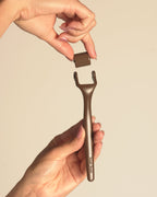 A close-up of a person's hands holding a metallic bronze facial roller tool and its matching cylindrical roller, set against a solid light beige background.