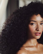 A close-up portrait of a woman with voluminous, tightly curled black hair, wearing a sheer nude strapless top. The background is a soft, out-of-focus neutral interior.
