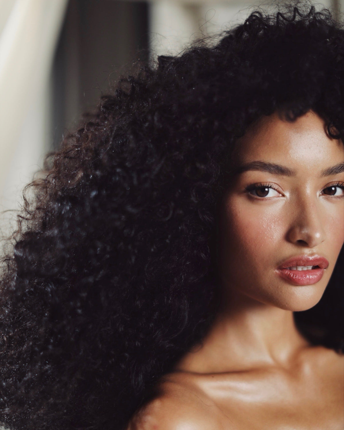 A close-up portrait of a woman with voluminous, tightly curled black hair, wearing a sheer nude strapless top. The background is a soft, out-of-focus neutral interior.