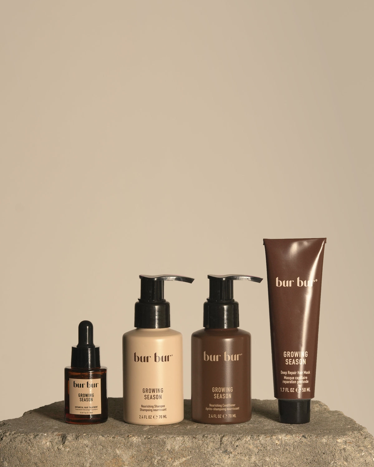 Four bur bur hair growth products arranged on a stone surface against a warm beige background: a small amber dropper bottle, a beige pump bottle, a brown pump bottle, and a brown squeeze tube.