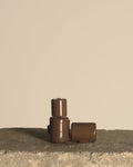 Three small brown cylindrical containers with embossed branding, arranged on a rough stone surface against a plain beige background.
