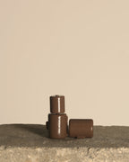 Three small brown cylindrical containers with embossed branding, arranged on a rough stone surface against a plain beige background.