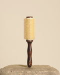 A round hairbrush with a dark brown wooden handle and a cream-colored bristle head, standing upright on a textured stone surface against a solid beige background.