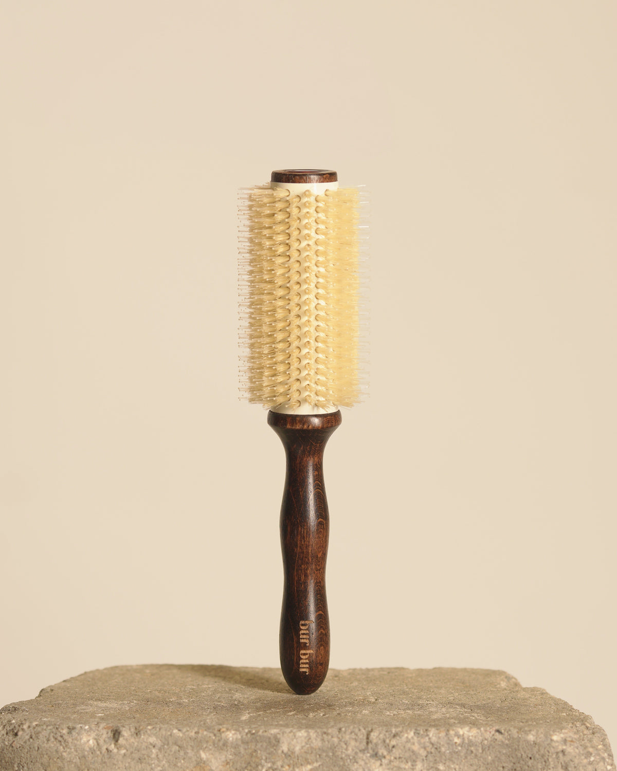 A round hairbrush with a dark brown wooden handle and a cream-colored bristle head, standing upright on a textured stone surface against a solid beige background.