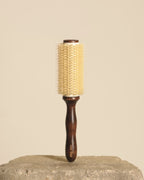 A round hairbrush with a dark brown wooden handle and a cream-colored bristle head, standing upright on a textured stone surface against a solid beige background.
