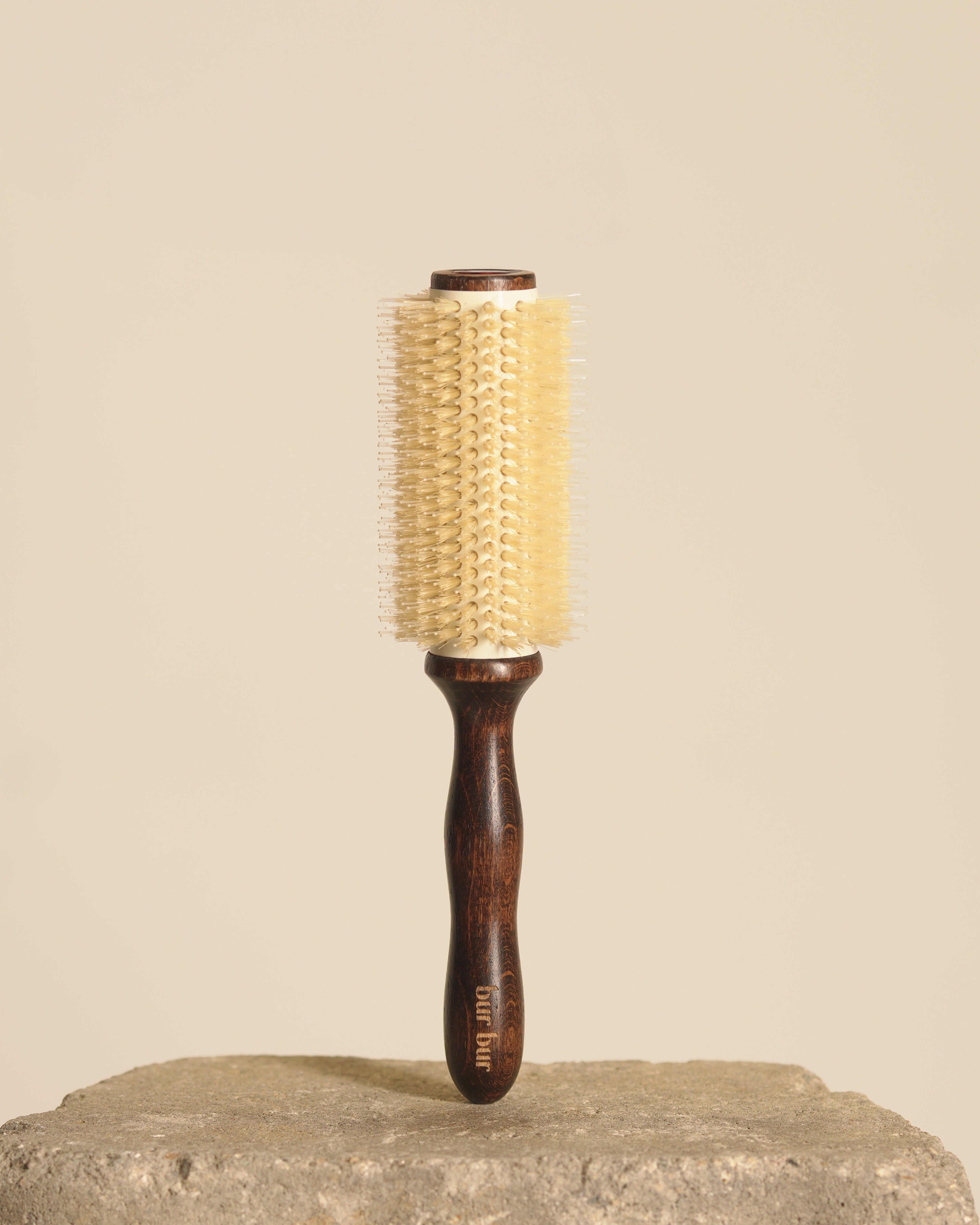 A round hairbrush with a dark brown wooden handle and a cream-colored bristle head, standing upright on a textured stone surface against a solid beige background.