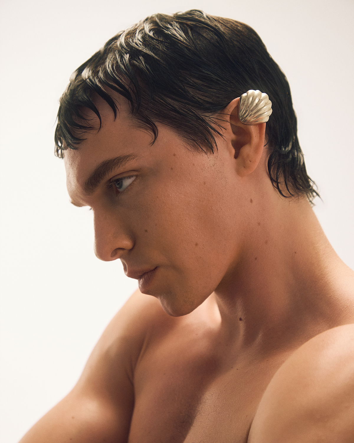 A close-up portrait of a shirtless man with wet dark hair wearing a white seashell in his ear, set against a plain light background.