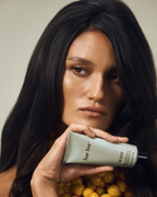 A close-up lifestyle shot of a model with long, straight black hair holding a tube of hair product. The background is filled with small yellow berries, and the product tube is labeled 'bar bur' and 'THE WISE'.