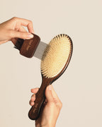 A close-up of a person's hands holding a wooden hairbrush with beige bristles and a smaller attached wooden comb tool against a plain warm beige background.