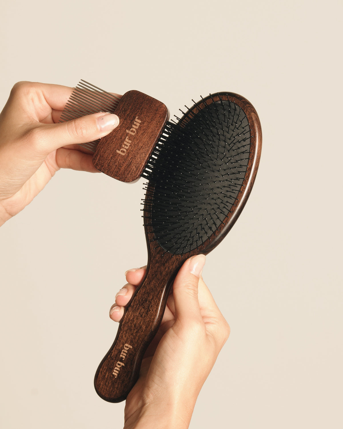 A person holds two wooden hairbrushes against a plain beige background. One larger oval brush has a dark wood handle and black bristles, while the smaller rectangular brush has a matching dark wood handle and fine bristles.
