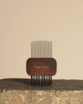 A wooden hair comb with fine metal teeth, standing upright on a rough stone surface against a plain beige background. The wooden handle is dark brown with the brand name 'bar bar' engraved on it.
