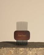 A wooden hair comb with fine metal teeth, standing upright on a rough stone surface against a plain beige background. The wooden handle is dark brown with the brand name 'bar bar' engraved on it.