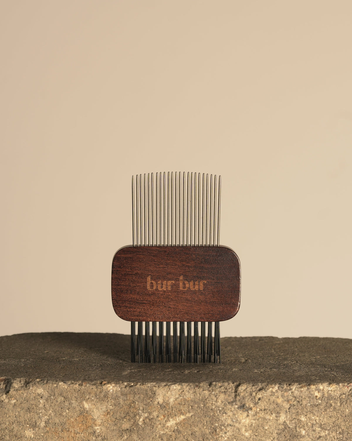 A wooden hair comb with fine metal teeth, standing upright on a rough stone surface against a plain beige background. The wooden handle is dark brown with the brand name 'bar bar' engraved on it.