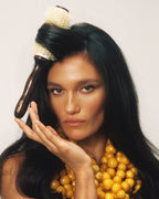 A woman with long black hair poses with a round hairbrush tucked into her hair, wearing a chunky yellow beaded necklace against a plain light background.